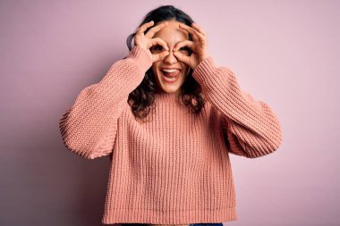 Kıvırcık saçlı genç bir kadın pembe arka planda günlük süveter giyiyor. Dürbün gibi güzel hareketler yapıyor. Gözleri parmaklarından bakıyor. Çılgın ifade.