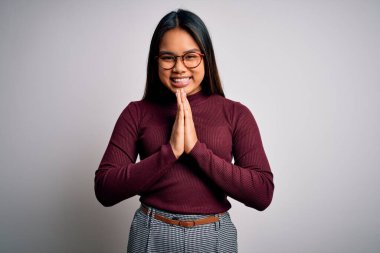 Güzel Asyalı iş kadını, beyaz arka plan üzerine günlük süveter ve gözlük takıp birlikte dua ederek af diliyor kendinden emin bir şekilde gülümsüyor..