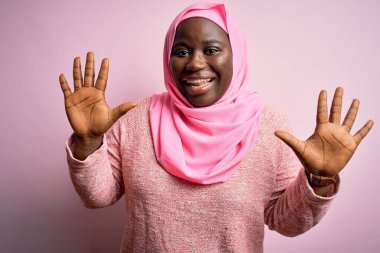 Genç Afro-Amerikan artı cüsseli kadın tecrit edilmiş pembe arka plan üzerine Müslüman başörtüsü takıyor ve parmaklarıyla on numarayı gösterirken kendinden emin ve mutlu gülümsüyor..