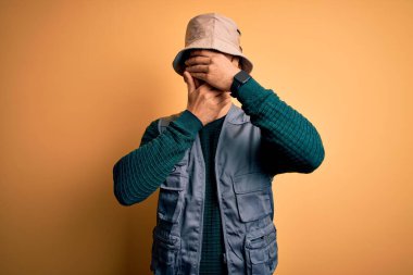 Tatilde sakallı yakışıklı turist sarı arka planda kaşif şapkası takıyor gözleri ve ağzı kapalı, şaşırmış ve şok olmuş. Duygularını gizliyor.