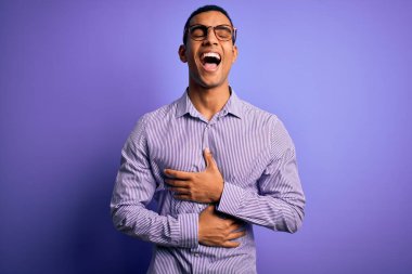 Yakışıklı Afro-Amerikalı adam mor arka planda çizgili gömlek ve gözlük takıyor. Gülümsüyor ve kahkahalar atıyor. Çünkü elleri vücutta olan komik bir şaka..