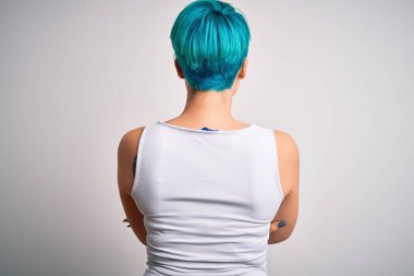 Mavi saçlı, beyaz arka planda sıradan tişörtler giyen, kollarını kavuşturup arkasını dönen genç ve güzel bir kadın.