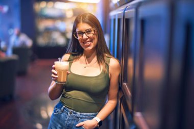 Mutlu ve kendinden emin gülümseyen genç ve güzel bir kadın. Yüzünde gülümsemeyle restoranda bir bardak kahve tutarken