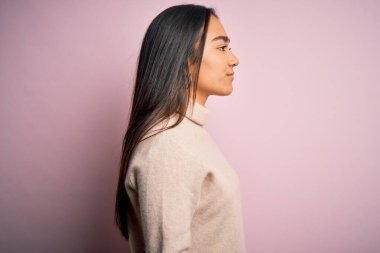 Genç, güzel Asyalı kadın pembe arka plan üzerine balıkçı yaka kazak giyiyor. Yan yan bakıyor, rahat poz veriyor, kendine güvenen bir gülüşü var..