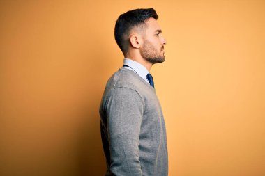 Zarif bir süveter ve kravat takan genç yakışıklı işadamı, sarı arka plana bakıyor, rahat bir görünüm sergiliyor, kendine güvenen bir gülümsemeyle doğal bir görünüm sergiliyor..