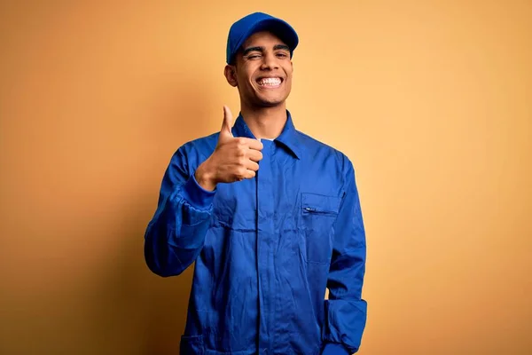 青い制服を着た若いアフリカ系アメリカ人の整備士が黄色の背景に帽子をかぶり 手で幸せな親指を立ててジェスチャーをします 成功を示すカメラを見て表現を承認する — ストック写真
