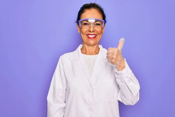 中年老年女科学家 身穿紫色背景的外套 戴着实验室眼镜 手拿着快乐的大拇指做着快乐的手势 在镜头前看着成功的表情 — 图库照片