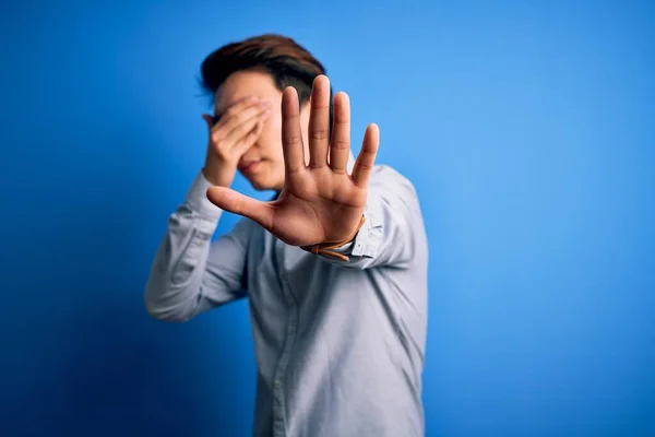 Young handsome chinese man wearing casual shirt standing over isolated blue background covering eyes with hands and doing stop gesture with sad and fear expression. Embarrassed and negative concept.