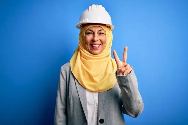 Middle age brunette architect woman wearing muslim traditional hijab and security helmet smiling with happy face winking at the camera doing victory sign. Number two.