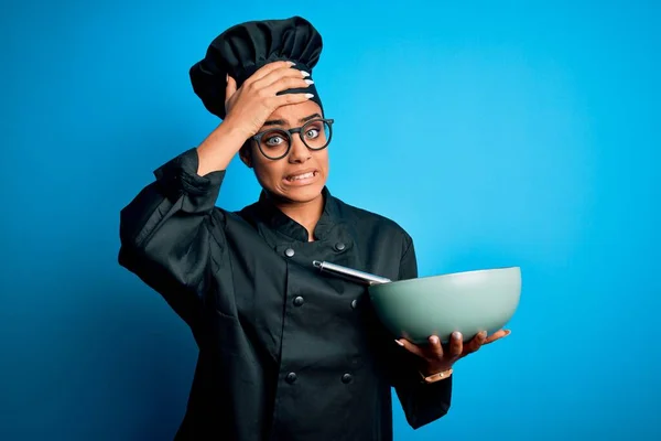 Young african american chef girl wearing cooker uniform and hat using whisk and bowl stressed with hand on head, shocked with shame and surprise face, angry and frustrated. Fear and upset for mistake.