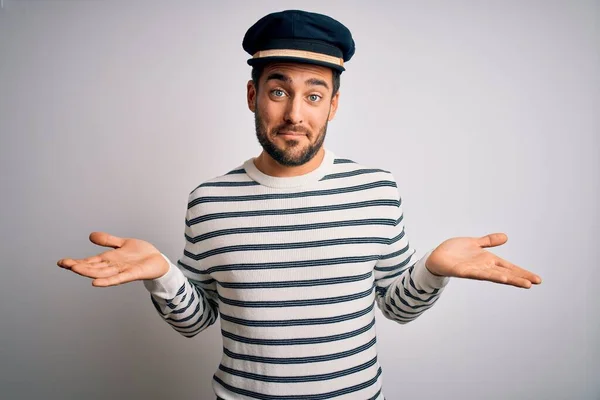 Young handsome sailor man with beard wearing navy striped uniform and captain hat clueless and confused expression with arms and hands raised. Doubt concept.