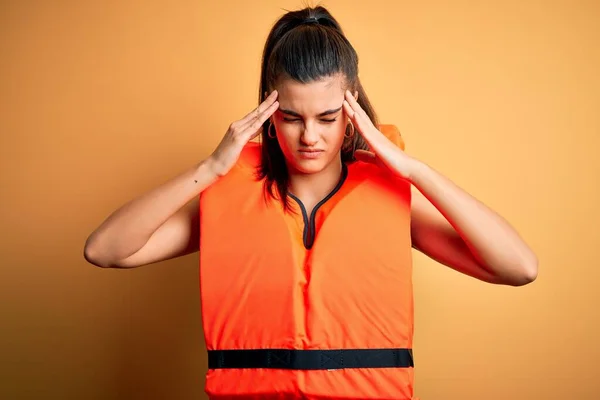 Young beautiful brunette woman wearing orange safe lifejacket over yellow background with hand on head for pain in head because stress. Suffering migraine.