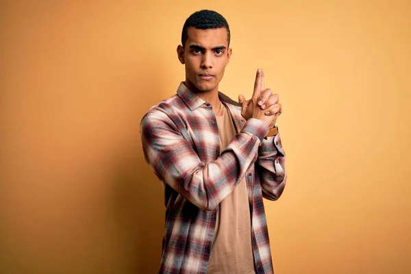 Young handsome african american man wearing casual shirt standing over yellow background Holding symbolic gun with hand gesture, playing killing shooting weapons, angry face