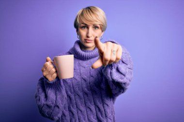 Kısa saçlı, kış süveteri giyen, kamerayı işaret eden bir fincan sıcak kahve içen genç sarışın kadın ve sen, el işareti, ön taraftan pozitif ve kendinden emin bir hareket.