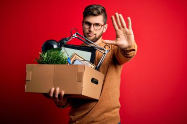 Sarışın, sakallı ve mavi gözlü genç adam elinde karton kutuyla elinde dur işareti, ciddi ve kendinden emin bir ifadeyle savunma hareketi yapıyor.