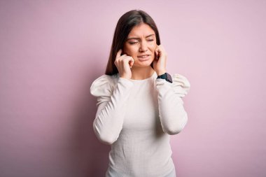 Mavi gözlü genç bir kadın pembe arka plan üzerine gündelik beyaz tişört giyip yüksek sesli müzik sesi için kulaklarını parmaklarıyla kapatıyor. Sağır kavramı.