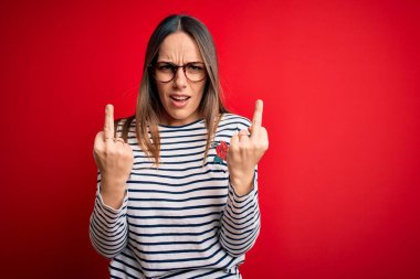 Young beautiful blonde woman with blue eyes wearing glasses standing over red background Showing middle finger doing fuck you bad expression, provocation and rude attitude. Screaming excited