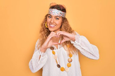 Genç, sarışın, mavi gözlü, güneş gözlüklü, aksesuar takan, aşkla gülümseyen, elleriyle kalp sembolü yapan güzel bir hippi kadın. Romantik konsept.