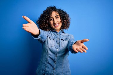 Genç ve güzel kıvırcık Arap kadın mavi arka planda dikilmiş kollarını açıp kameraya gülümseyen günlük kot gömlek giyiyor. Mutluluğu kucaklayan neşeli bir ifade..