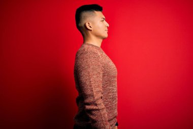 Kırmızı arka planda dikilen, rahat bir görünüm veren, kendine güvenen gülümseyen, gündelik süveter giyen genç, yakışıklı Latin adam..