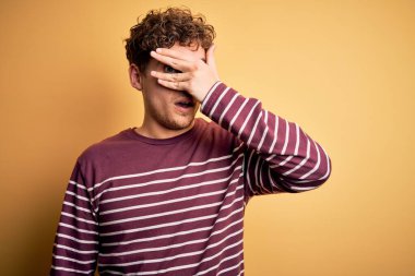 Sarışın, kıvırcık saçlı, sıradan çizgili süveter giyen, yüzünde ve gözlerinde utangaç ifadesiyle parmaklarının arasından bakan genç, yakışıklı bir adam..