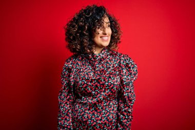 Genç, güzel, kıvırcık bir Arap kadın. Çiçekli bir elbise giyiyor. Kırmızı arka planda duruyor. Kendinden emin gülen.