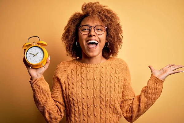 Young beautiful African American afro woman with curly hair holding vintage alarm clock very happy and excited, winner expression celebrating victory screaming with big smile and raised hands