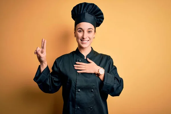 Young beautiful chef woman wearing cooker uniform and hat standing over yellow background smiling swearing with hand on chest and fingers up, making a loyalty promise oath