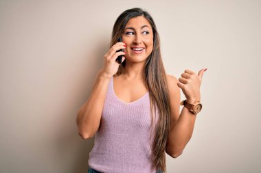 Genç esmer kadın beyaz arka planda akıllı telefondan konuşuyor ve gülümseyen bir gülümsemeyle baş parmağıyla onu işaret ediyor.