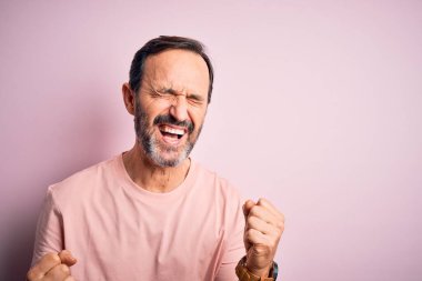 İzole edilmiş pembe arka plan üzerinde dikilen orta yaşlı, ağarmış bir adam mutlu ve heyecanlı bir şekilde kollarını kaldırıp, gülümseyerek ve başarı için çığlık atarak zafer jesti yapıyor. Kutlama konsepti.