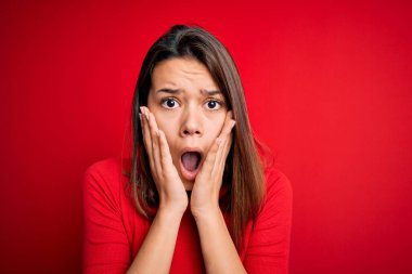 Genç ve güzel esmer kız, izole edilmiş kırmızı arka plan üzerinde gündelik tişört giyiyor. Korkmuş ve şok olmuş, şaşırmış ve şaşırmış bir halde.