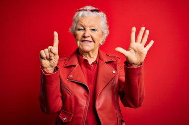 Son sınıf güzeli, gri saçlı, günlük kırmızı ceket giyen, güneş gözlüklü, altı numaralı parmaklarıyla işaret eden, kendinden emin ve mutlu bir şekilde gülümseyen kadın..