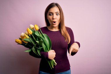 Sarı lalelerden oluşan romantik buketi pembe arka planda tutan genç sarışın kadın parmaklarını göstererek reklamını yapıyor, şaşırmış yüzünü ve ağzını açıyor.