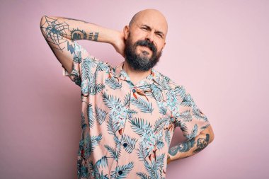 Sakallı ve dövmeli yakışıklı bir adam pembe arka planda sıradan bir çiçek gömleği giyiyor boynu ağrıyor, eliyle boynuna dokunuyor, kas ağrısı çekiyor.
