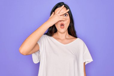 Genç, güzel, esmer bir kadın, mor arka plan üzerine gündelik beyaz tişört giyiyor. Şok olmuş bir şekilde yüzünü ve gözlerini kapıyor, utanmış bir ifadeyle parmaklarından bakıyor..