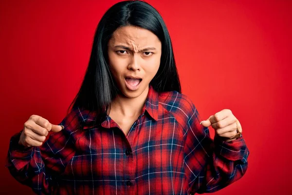 Young beautiful chinese woman wearing casual shirt over isolated red background angry and mad raising fists frustrated and furious while shouting with anger. Rage and aggressive concept.