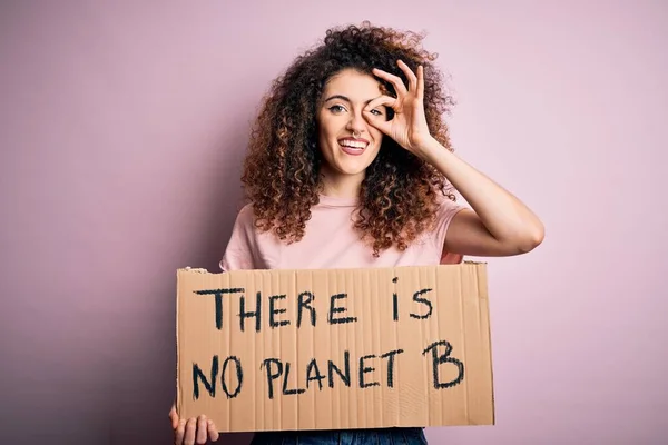 Young beautiful activist woman with curly hair and piercing protesting asking for change planet with happy face smiling doing ok sign with hand on eye looking through fingers
