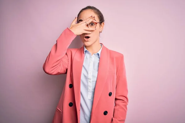 Young beautiful businesswoman wearing jacket and glasses over isolated pink background peeking in shock covering face and eyes with hand, looking through fingers with embarrassed expression.