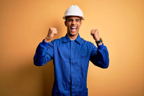 Young handsome african american worker man wearing blue uniform and security helmet celebrating surprised and amazed for success with arms raised and open eyes. Winner concept.