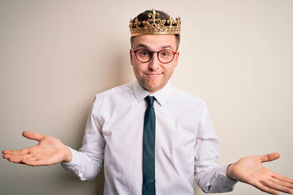 Молодой красивый кавказский бизнесмен, одетый в золотую корону на изолированном фоне, невежественное и запутанное выражение лица с поднятыми руками. Концепция сомнений
.