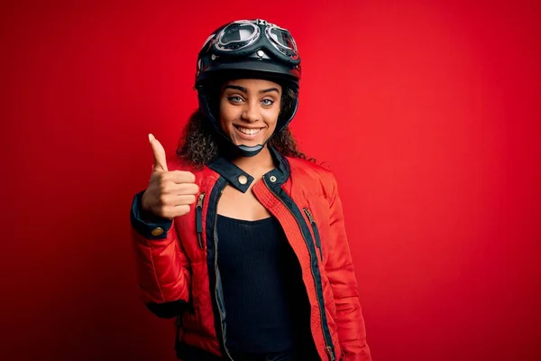 赤い背景の上に元ヘルメットと眼鏡をかけた若いアフリカ系アメリカ人のオートバイ愛好家の女の子が手で幸せな親指をジェスチャーしています 成功を示すカメラを見て表現を承認する — ストック写真