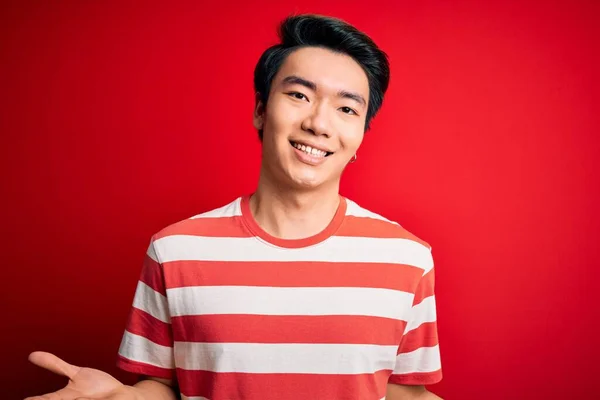 若いですハンサムな中国人男性身に着けているカジュアルストライプTシャツ上に立つ赤い背景笑顔陽気にオープン腕として優しい歓迎 正と自信を持って挨拶 — ストック写真