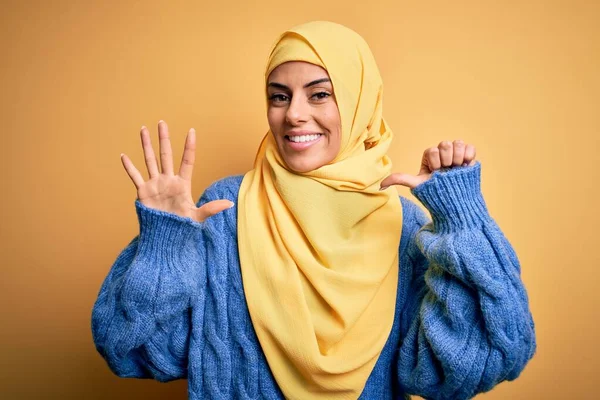 Young beautiful brunette muslim woman wearing arab hijab over isolated yellow background showing and pointing up with fingers number six while smiling confident and happy.