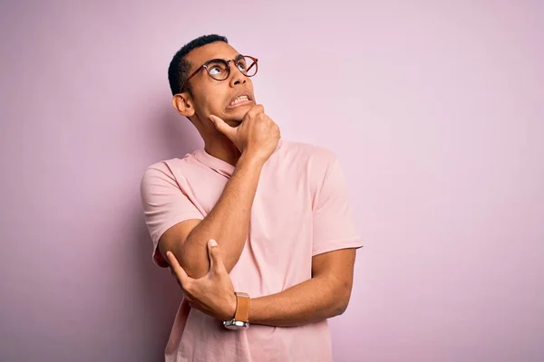 Handsome african american man wearing casual t-shirt and glasses over pink background Thinking worried about a question, concerned and nervous with hand on chin
