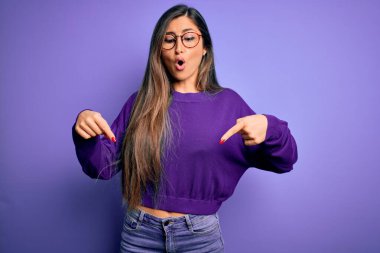 Genç, güzel, zeki, gözlüklü, mor tenha arka plan gözlüklü bir kadın. İlan veren parmaklarıyla işaret ediyor, şaşırmış yüzünü ve ağzını açıyor.