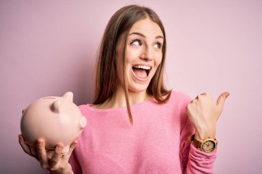 Genç, güzel, kızıl saçlı bir kadın kumbarayı soyulmuş pembe arka planda tutuyor ve gülümseyen bir gülümsemeyle baş parmağıyla işaret ediyor.