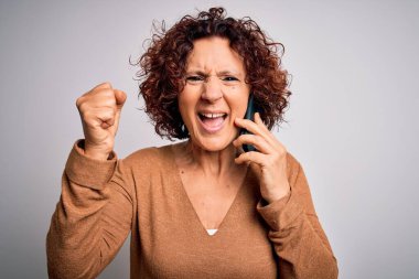 Orta yaşlı kıvırcık kadın beyaz arka planda telefonla konuşuyor. Gururla bağırıyor, zaferi ve başarıyı kutluyor. Çok heyecanlı, coşkulu bir duygu.