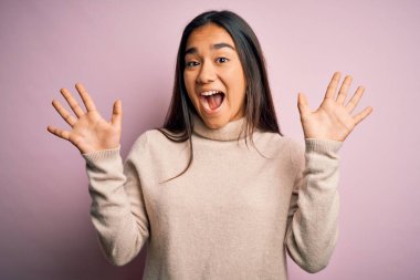 Genç, güzel Asyalı kadın pembe arka planda balıkçı yaka kazak giyiyor ve parmaklarını on numarayı gösterirken kendinden emin ve mutlu bir şekilde gülümsüyor..