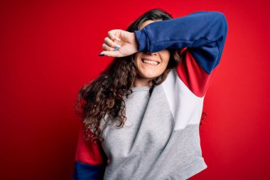 Kıvırcık saçlı genç bir kadın izole edilmiş kırmızı arka plan üzerine günlük eşofman giyip kolları güler yüzlü ve komik bir şekilde gözlerini kapatıyor. Kör kavram.