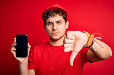 Kıvırcık saçlı, şapkalı, kırmızı arka planda akıllı telefon tutan, sinirli yüzlü, olumsuz işaretli, baş parmakları aşağıda, reddedilme konsepti olan genç sarışın teslimatçı.
